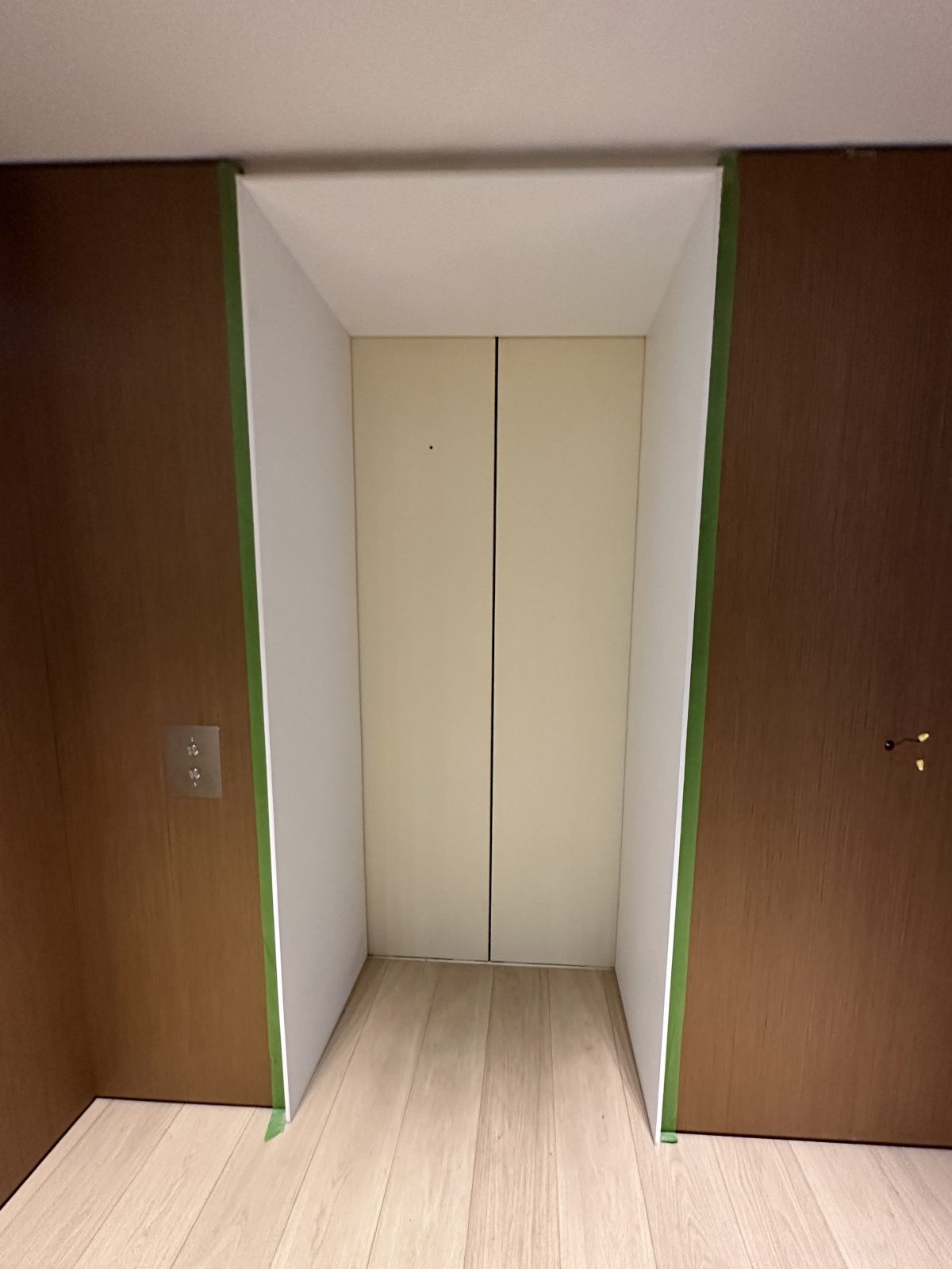 A timber lined residential elevator entrance with cream painted doors and a white reveal, green masking tape run down both junctions