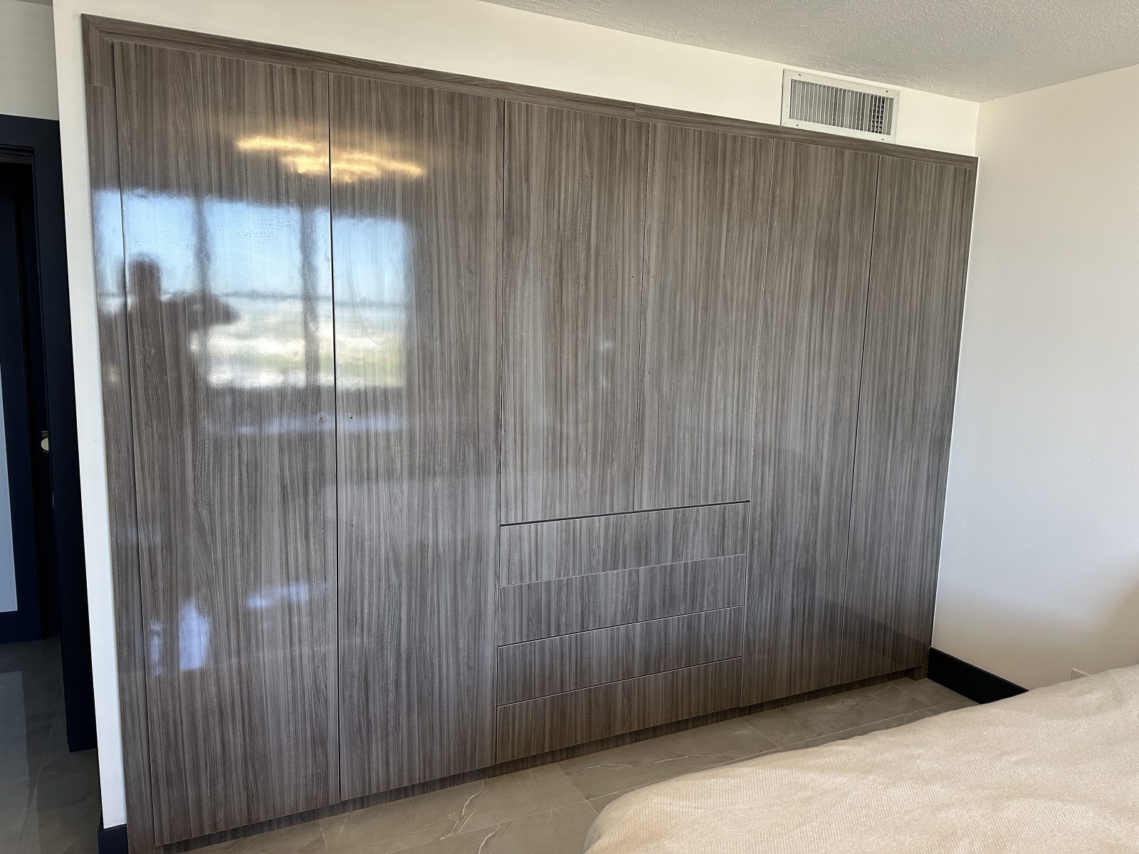 A wall of fitted wardrobes in high gloss grey woodgrain, reflecting the bedroom window across every door
